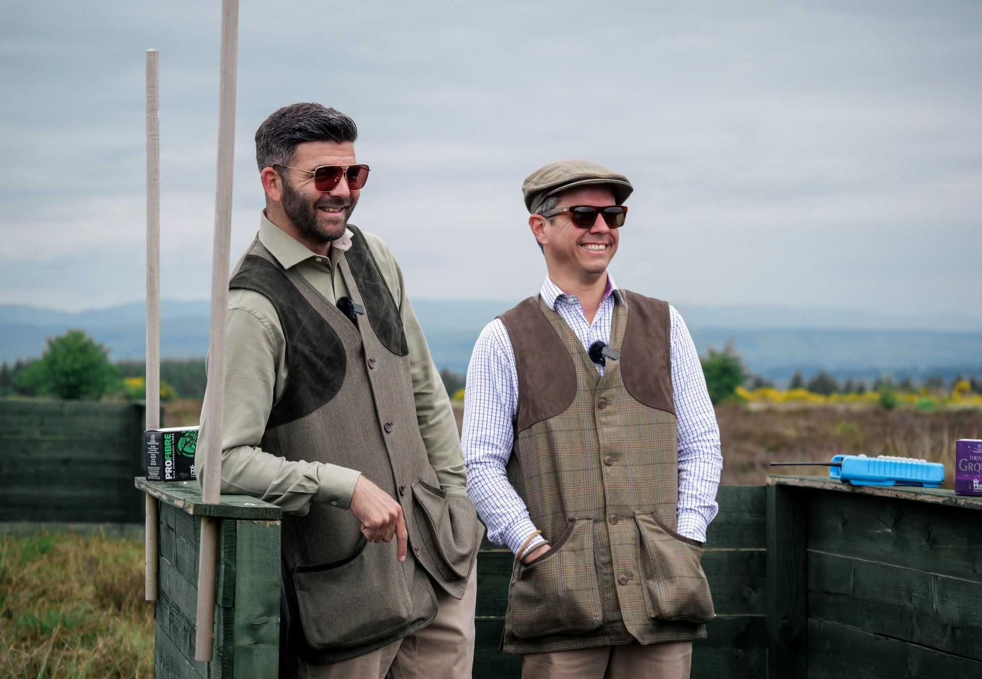 Two men in vests and caps smile at a shooting range. One leans against a wooden structure, the other stands.