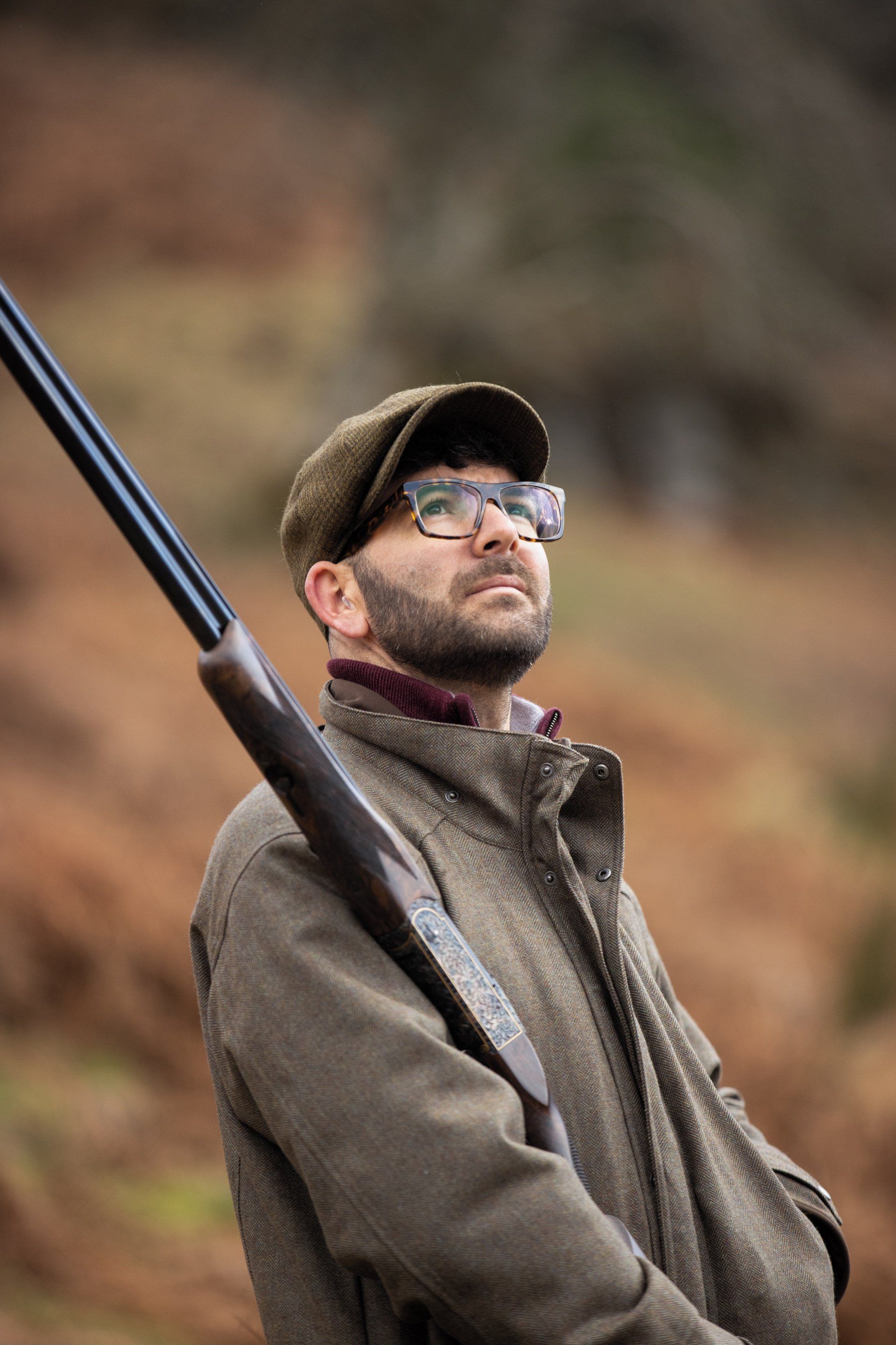 Man in tweed jacket and cap, holding a shotgun, looking up. Forest background.