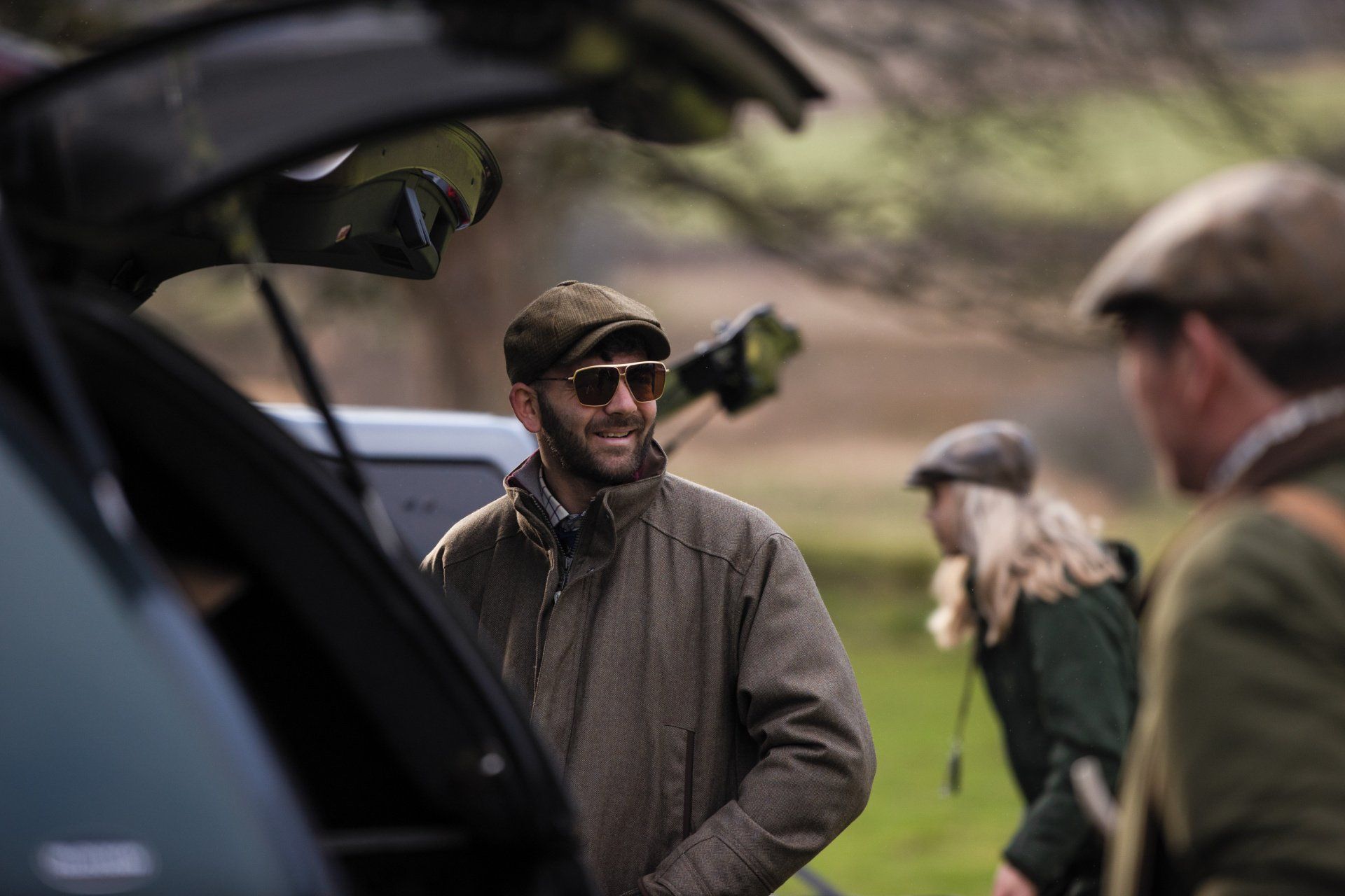 Man in tweed jacket and cap talks near an open car, with others in the background outdoors.