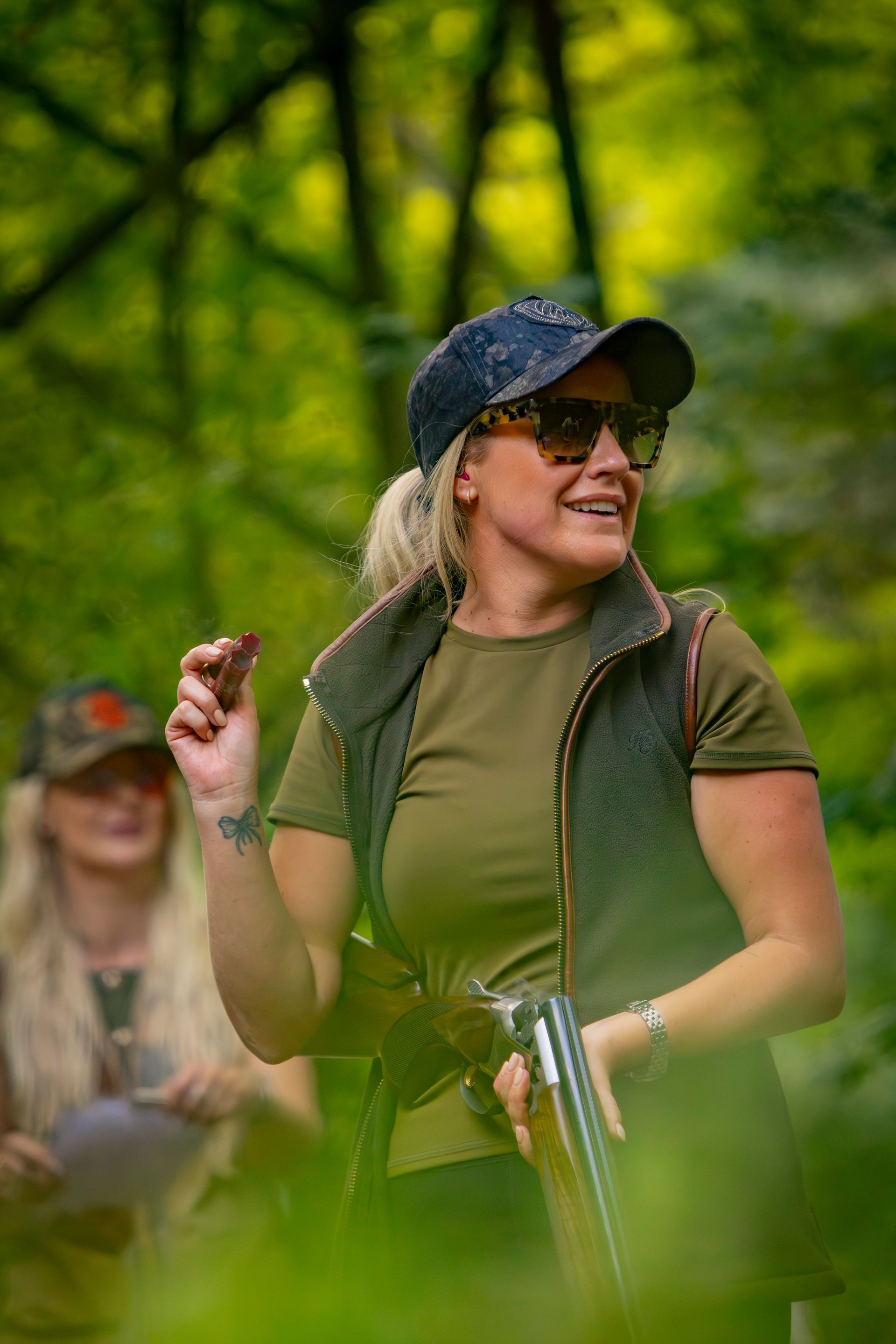 Woman with shotgun and cap, smiles outdoors. Holding something, another person in the blurred background.