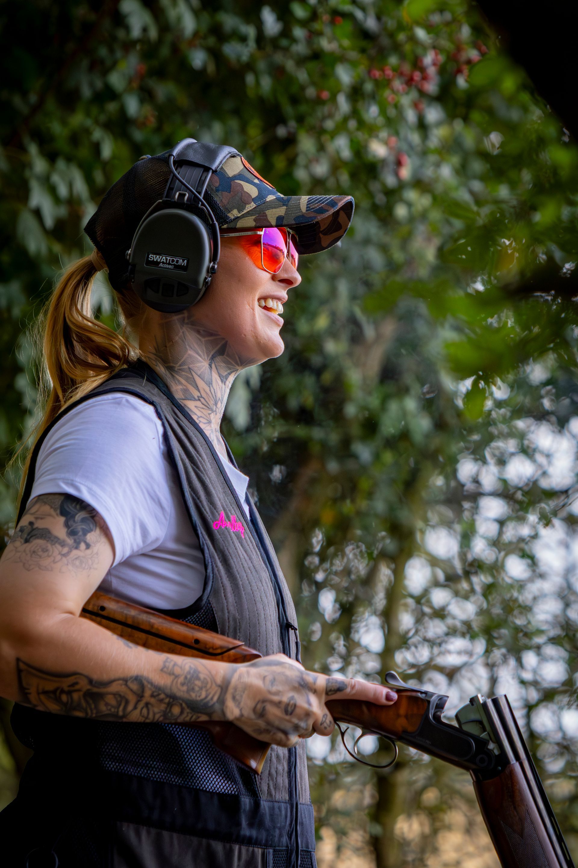 Woman with tattoos, shooting a shotgun, wearing safety glasses and ear protection, smiling, outdoors.