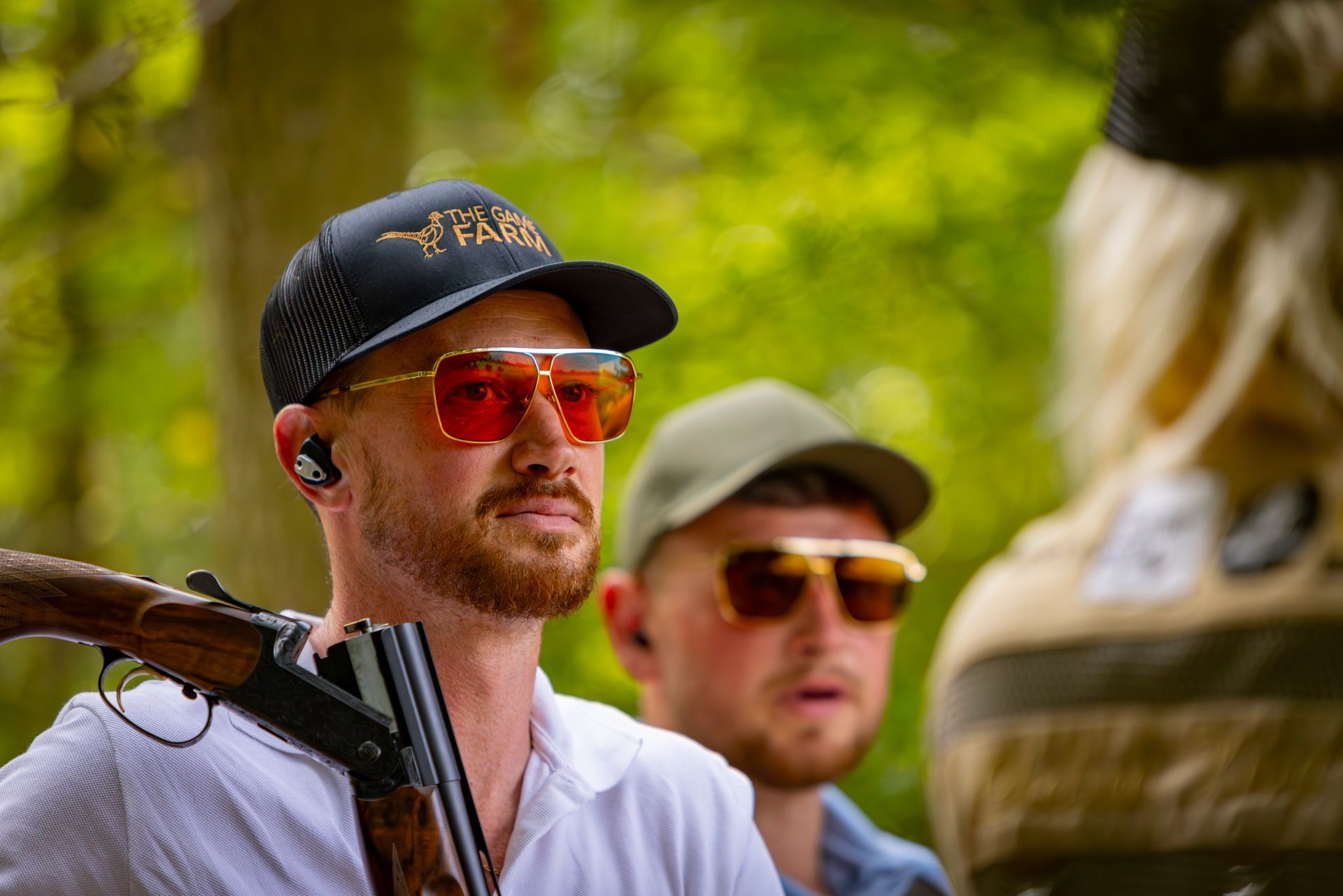 Man with red-tinted sunglasses holding a shotgun, looking to the side. Another man in the background. Outdoor setting.