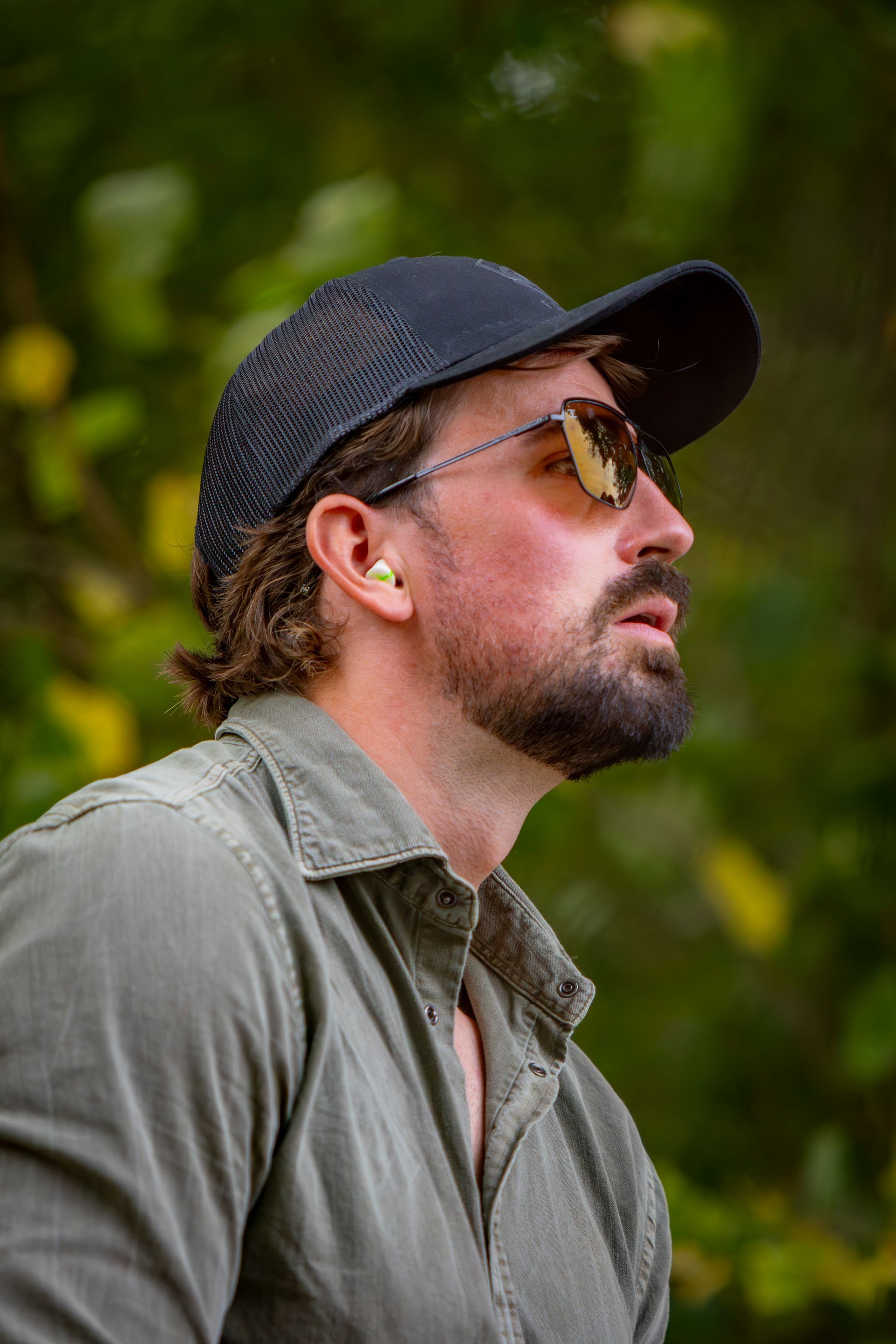 Man in cap and sunglasses, looking upward with a serious expression, wearing an olive shirt.
