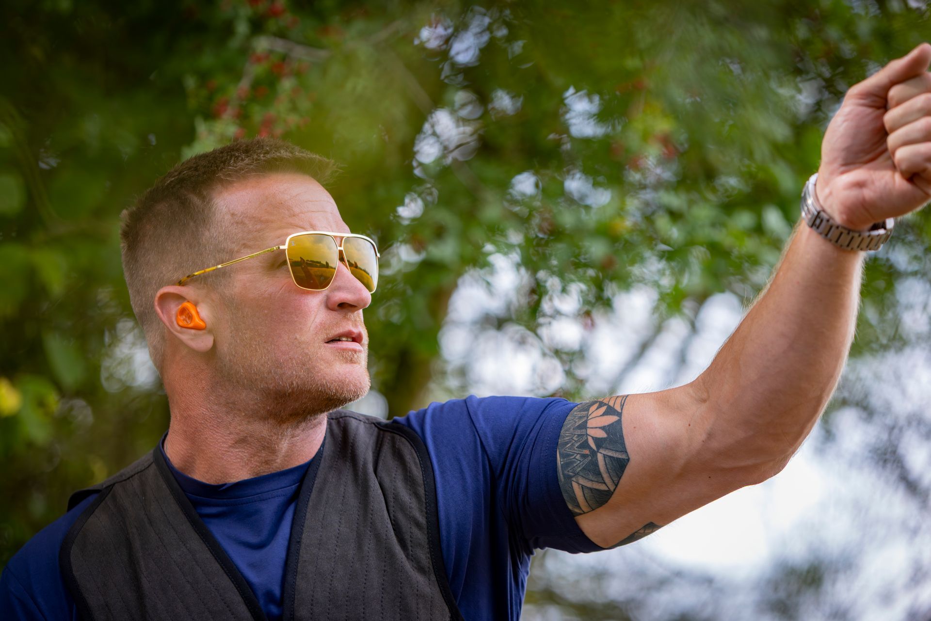 Man wearing sunglasses and earplugs raises arm, possibly shooting, in a green outdoor setting.