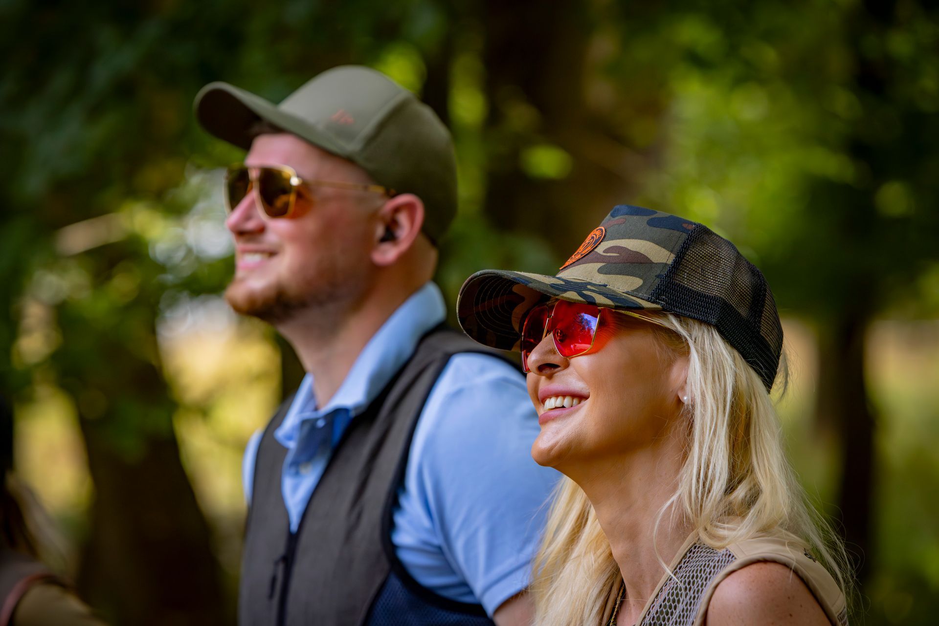 Two people with caps and sunglasses smile outside; man wears a vest.