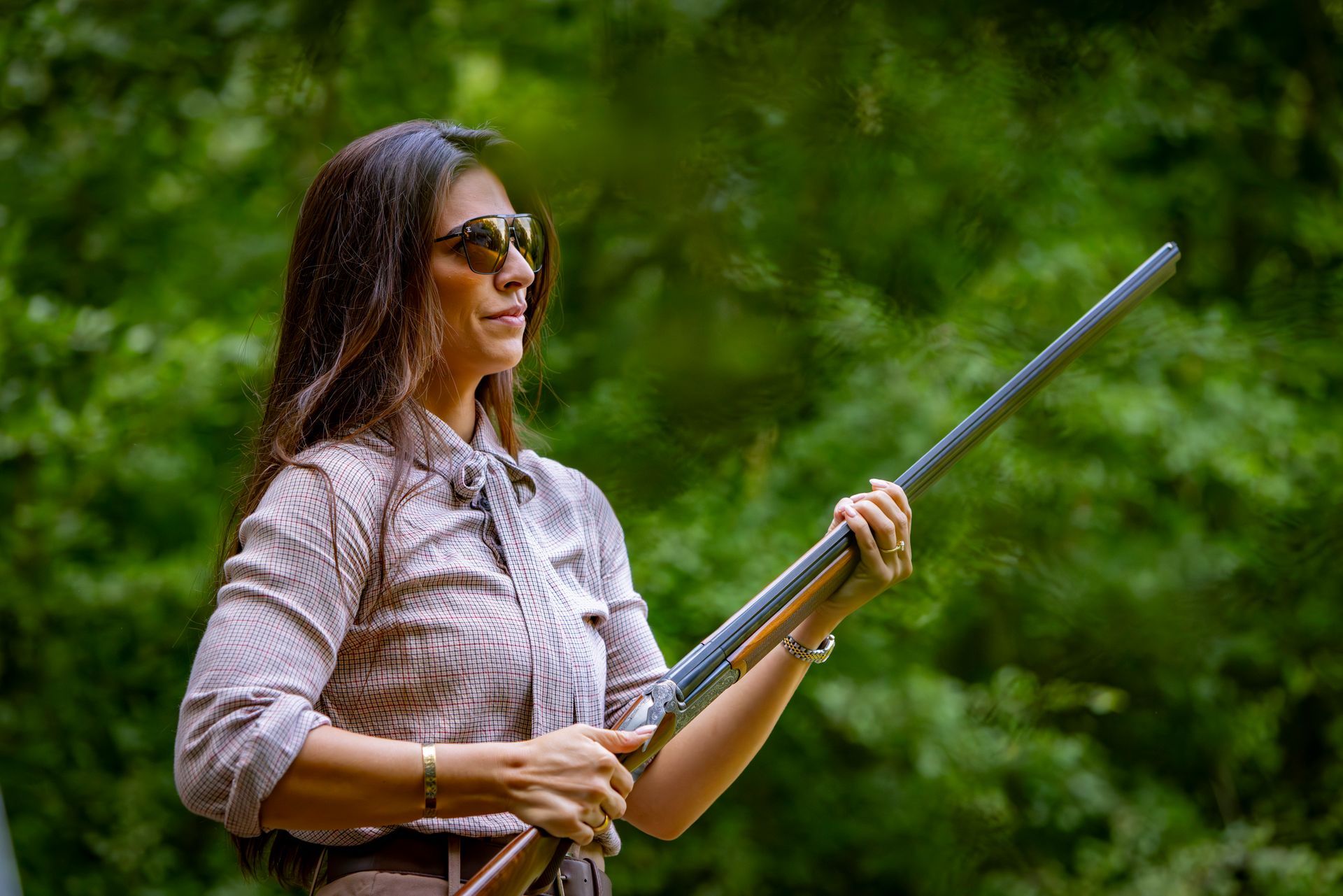Woman in sunglasses holding a long gun, outdoors, green foliage background.