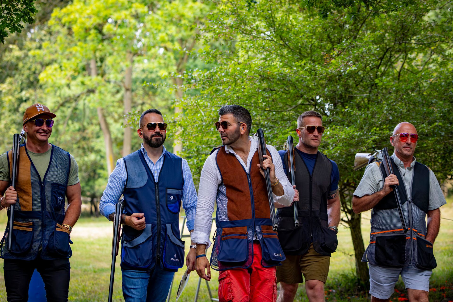 Five people in shooting vests, holding shotguns, walking outdoors.