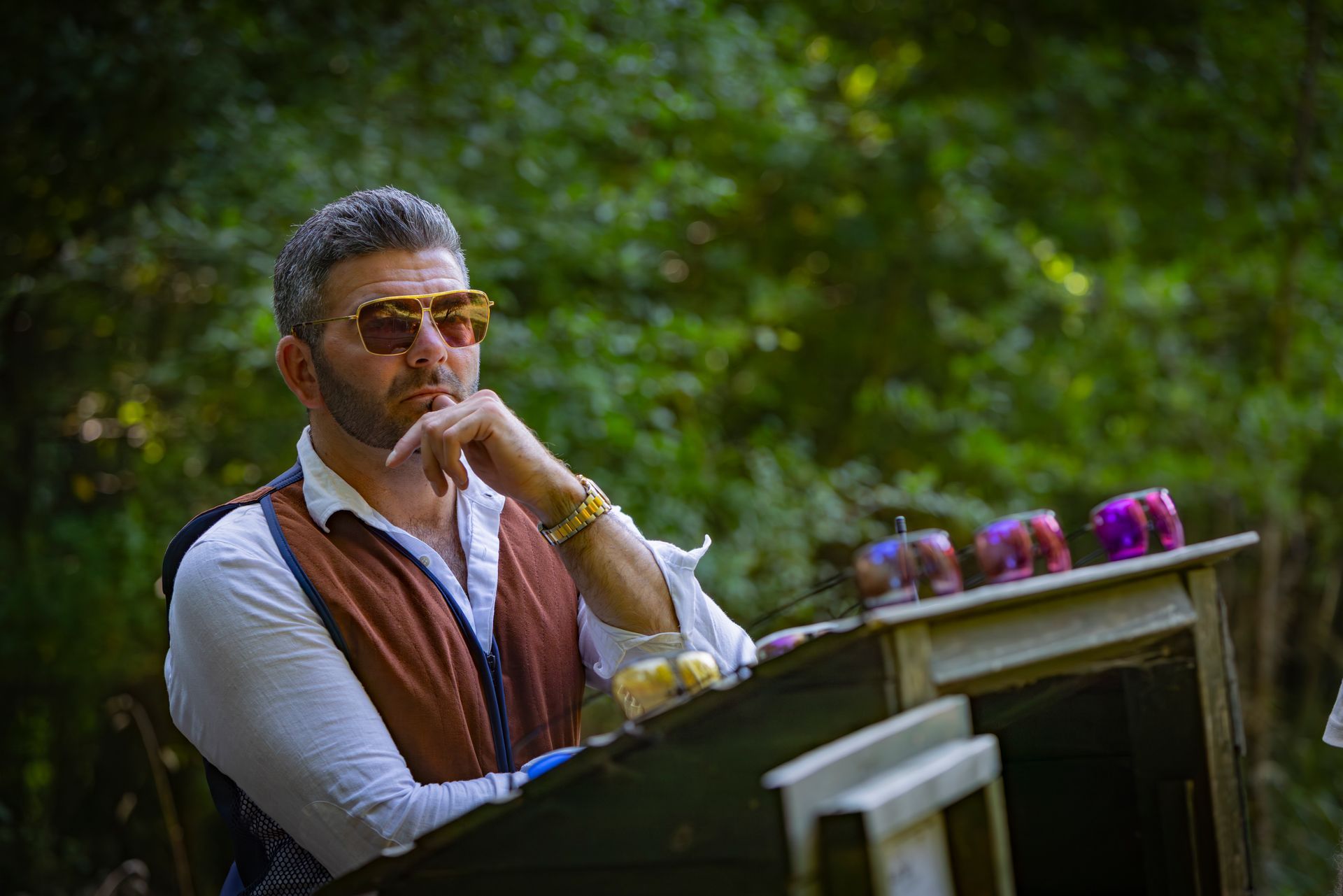 Man wearing sunglasses and vest, resting chin on hand, looking off, near a table with candles in a forest.