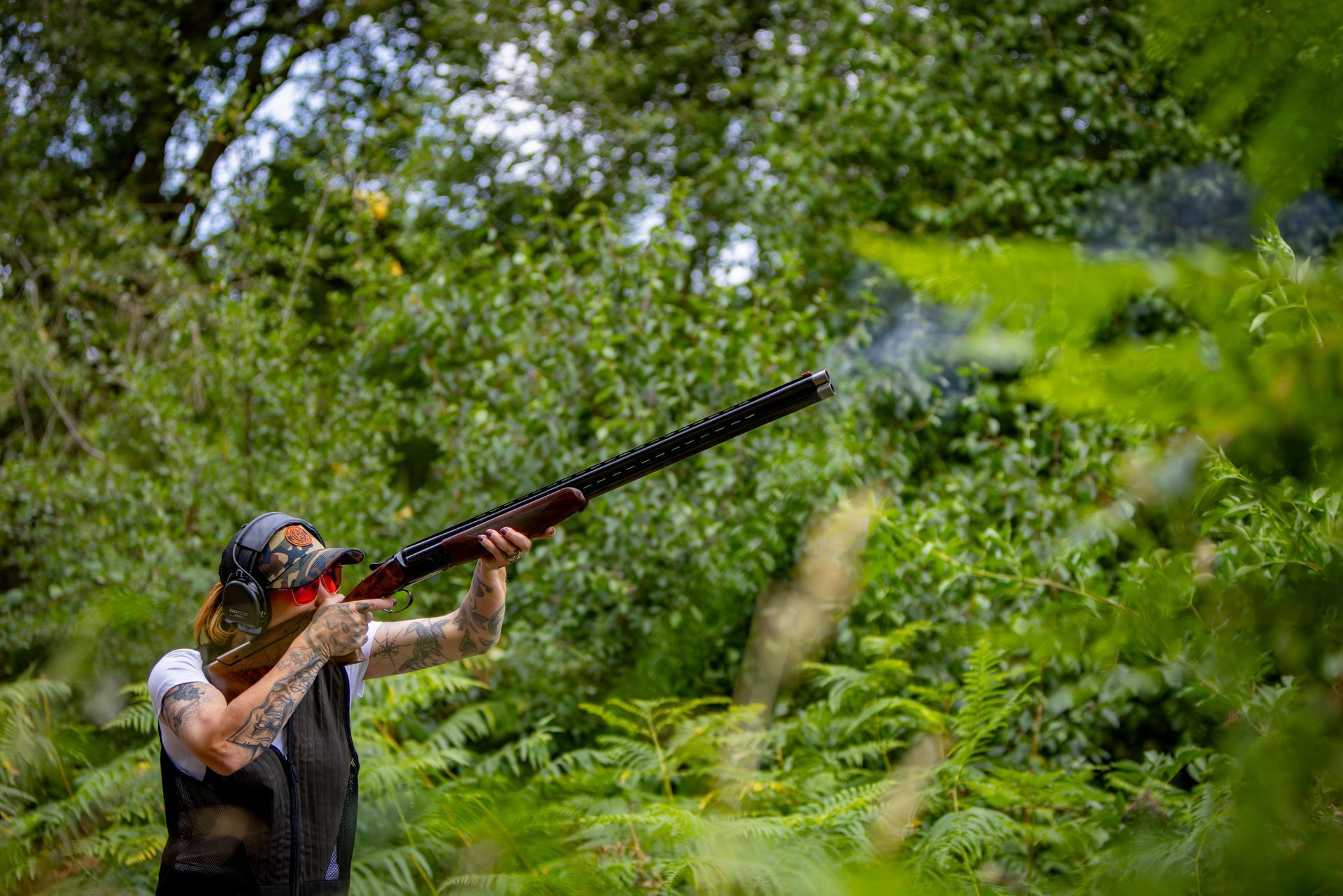 Person shooting a shotgun in a forest setting; smoke visible from the barrel.