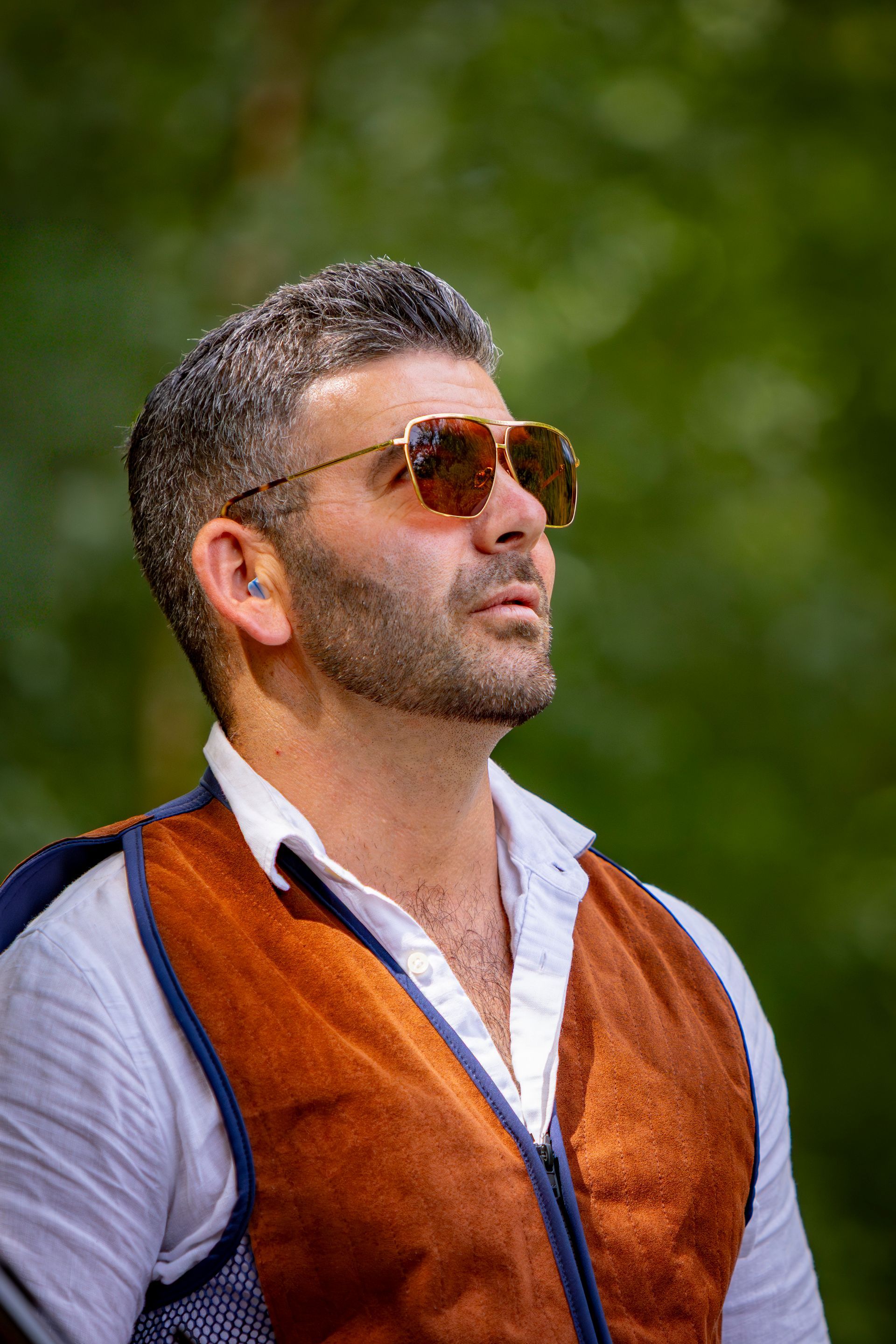 Man wearing sunglasses, brown vest, and white shirt, looking upward against a blurred green background.