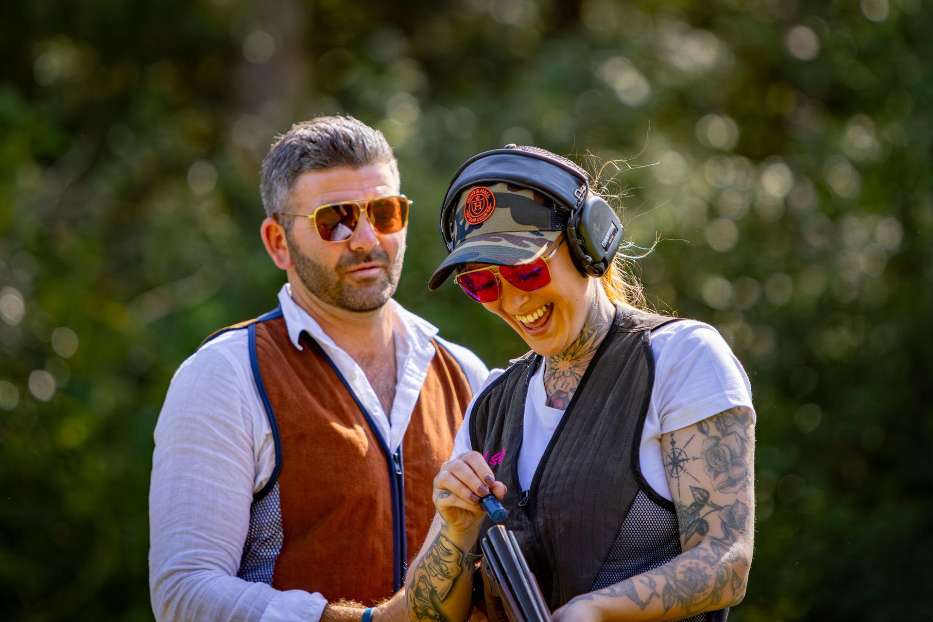 Man and woman at a shooting range. Woman laughs, holding a shotgun; both wear eye/ear protection.