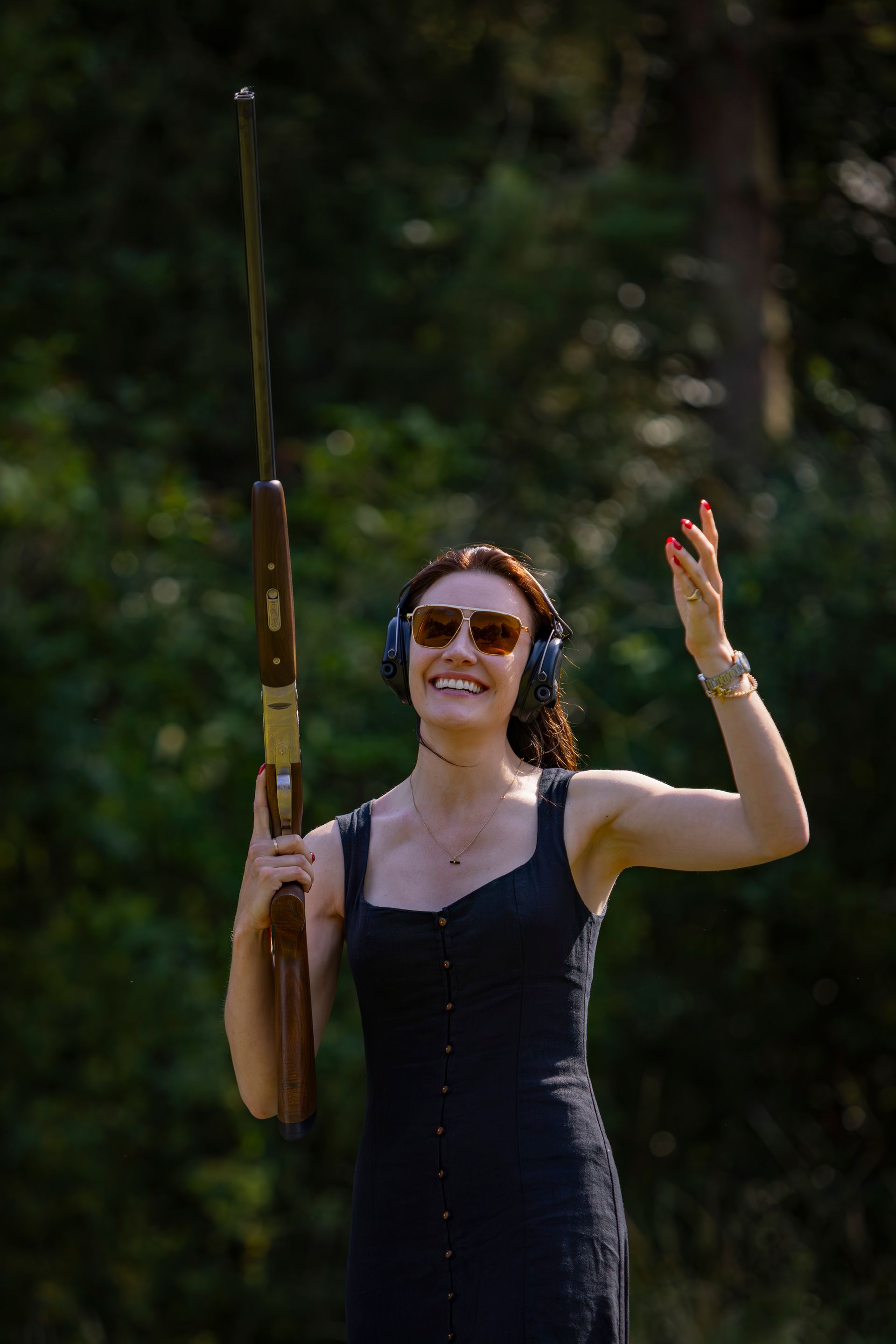 Woman in sunglasses, ear protection, and black dress, holding a rifle and smiling in an outdoor setting.