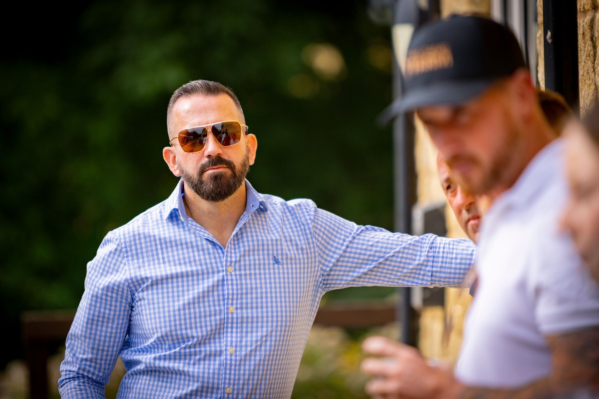Man with beard and sunglasses, leaning, blue checked shirt, observing others outdoors.