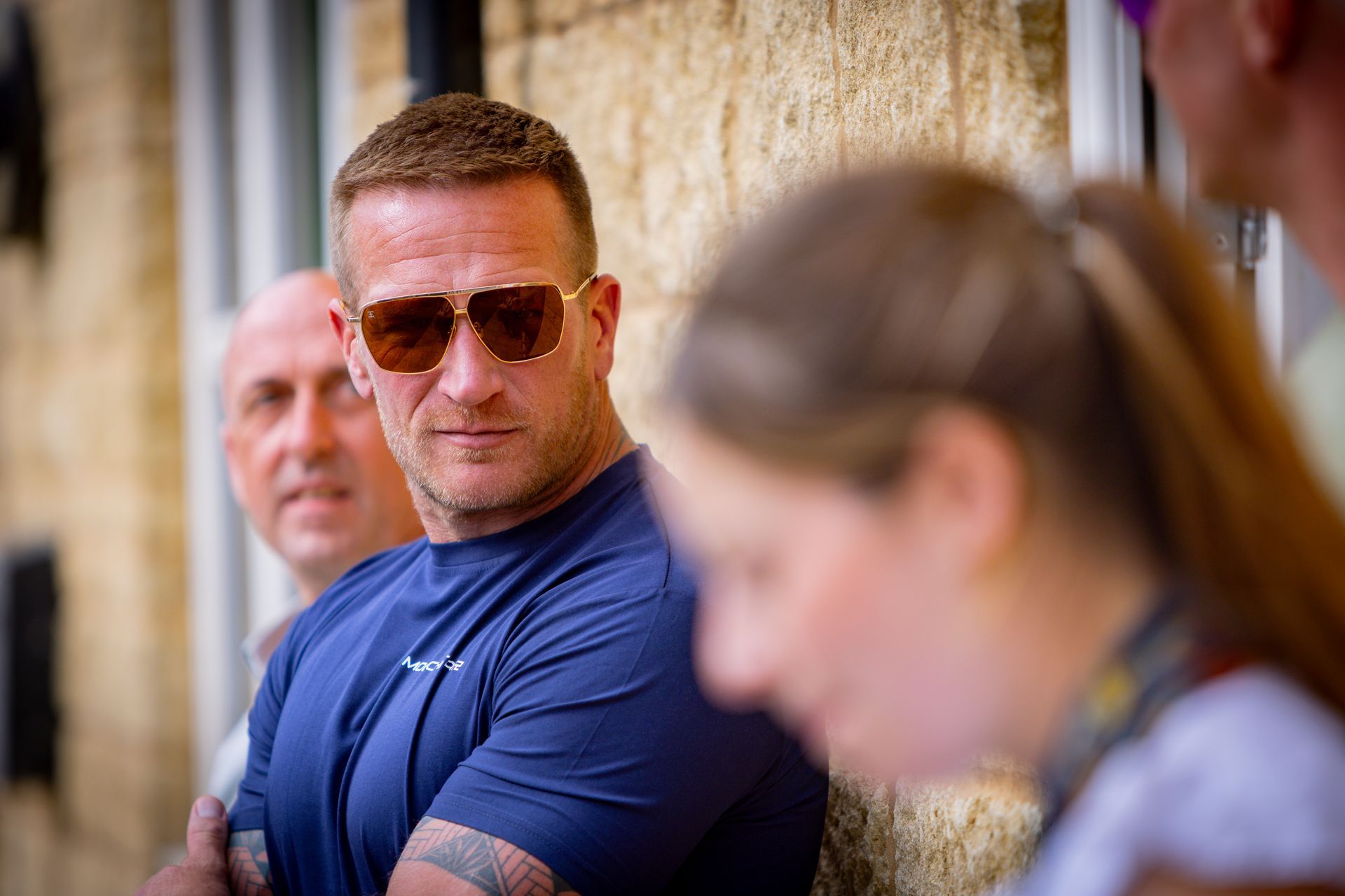 Man in sunglasses, blue shirt, arms crossed, looking intently. People in background.