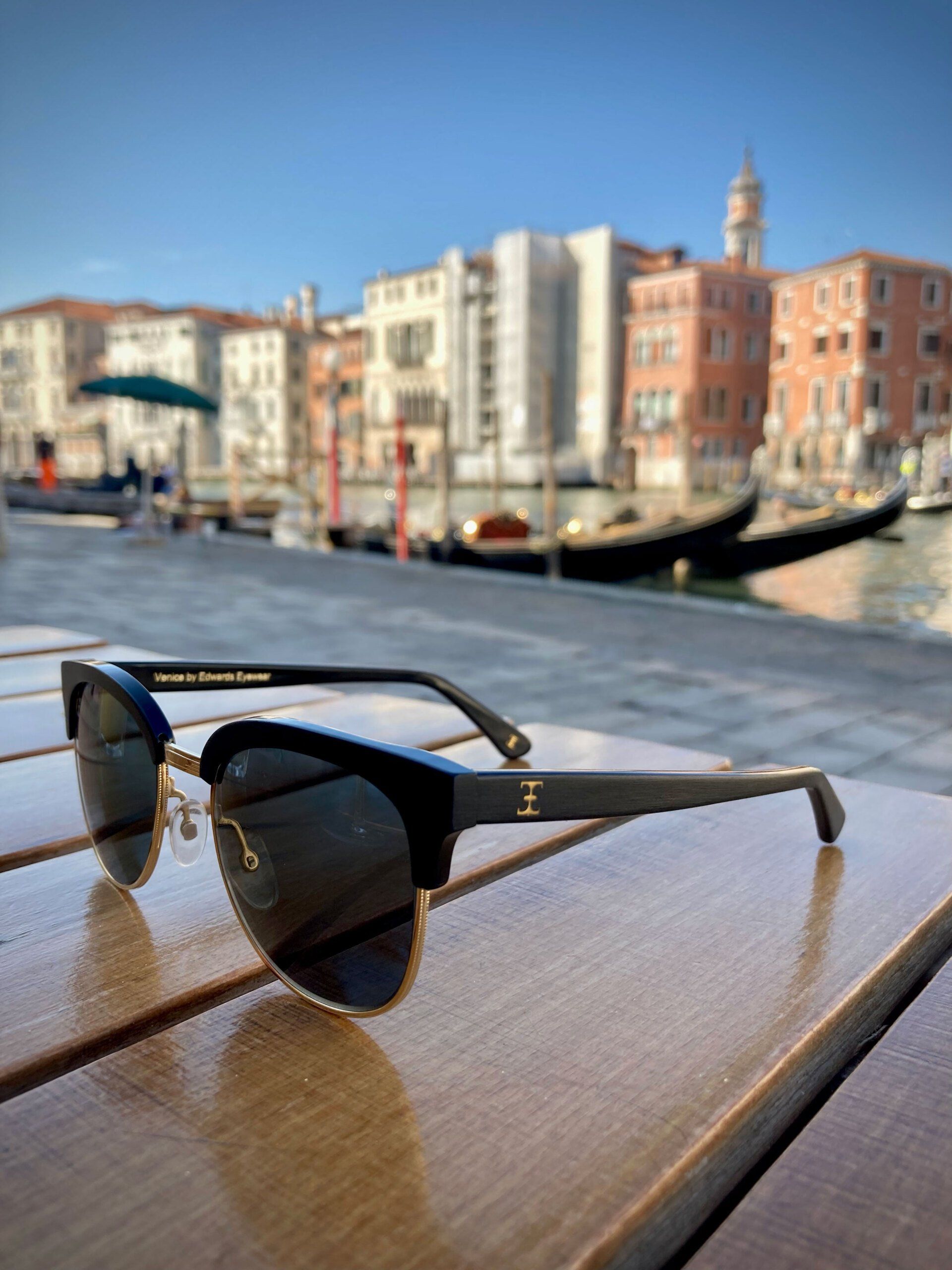 Sunglasses on a wooden surface with a Venetian canal and buildings in the blurred background.