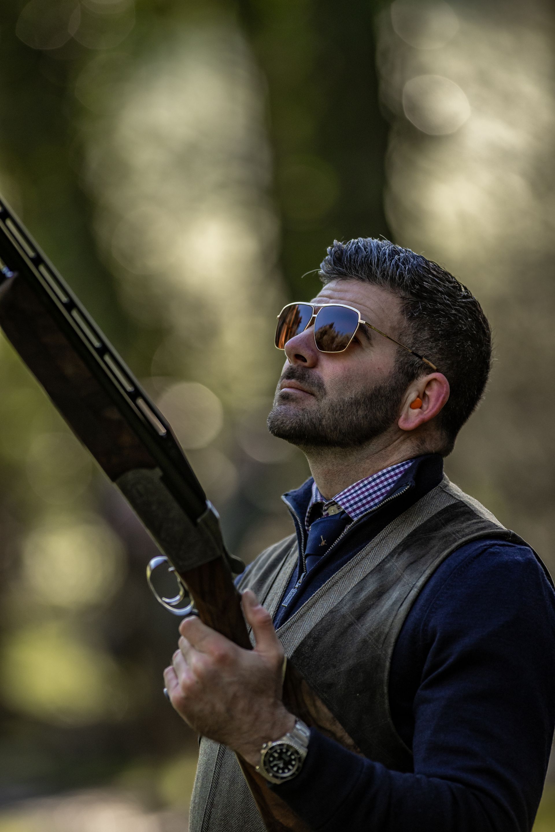 Man holding a shotgun, looking upward, wearing sunglasses, vest, and watch, outdoors.