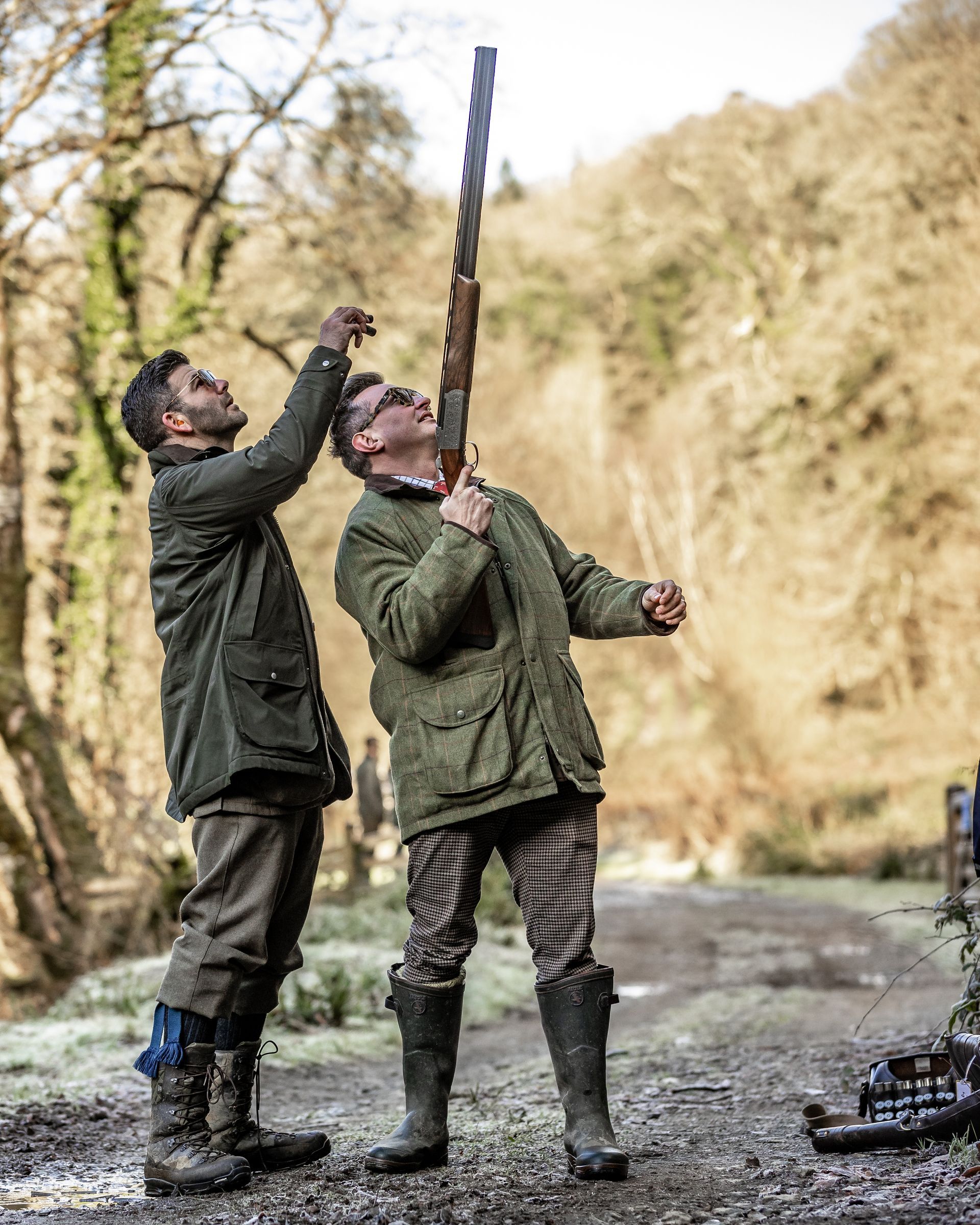 Two men, in green jackets and boots, inspecting a tall rifle outdoors.