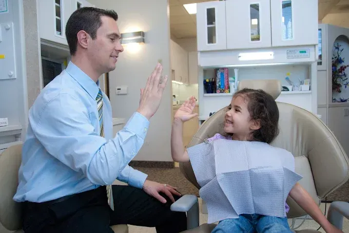 Dentist giving a high five to a young patient in a dental chair, smiling, light blue shirt.