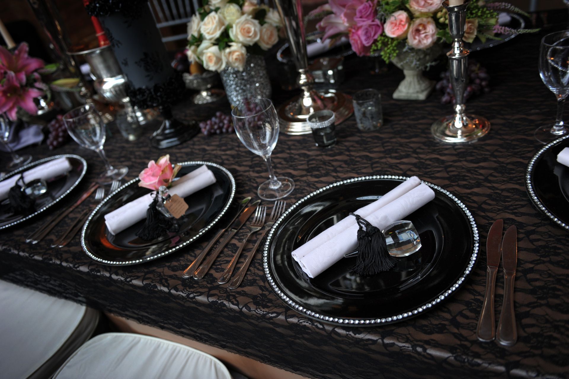 Elegant dinner table set with black plates, rolled napkins, candle holders, and pink floral centerpieces