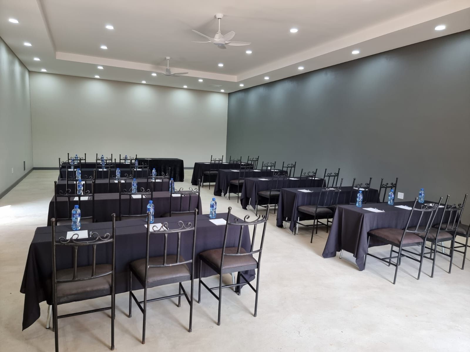 Empty banquet room with rows of black-draped tables and chairs under ceiling lights and fans