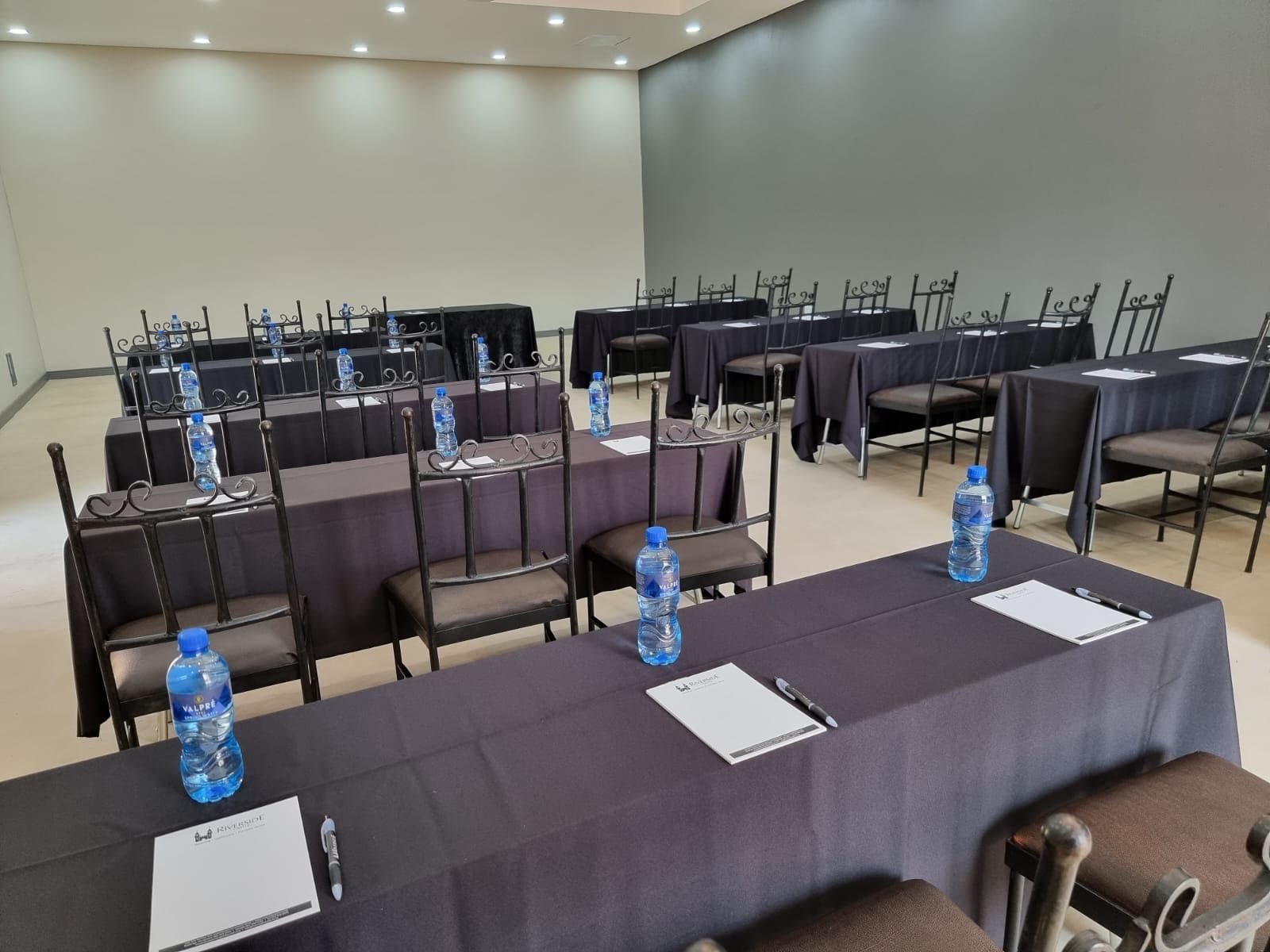 Empty conference room with rows of black tables and chairs, blue water bottles, and notepads arranged for a meeting