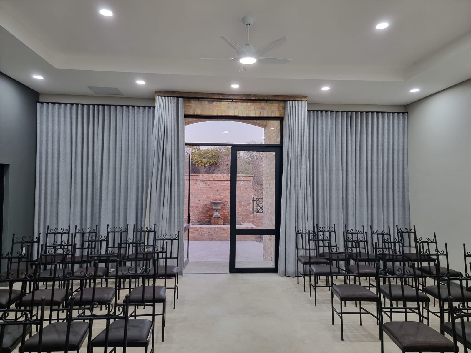 Empty indoor hall with rows of black chairs and gray curtains facing a glass door and brick courtyard