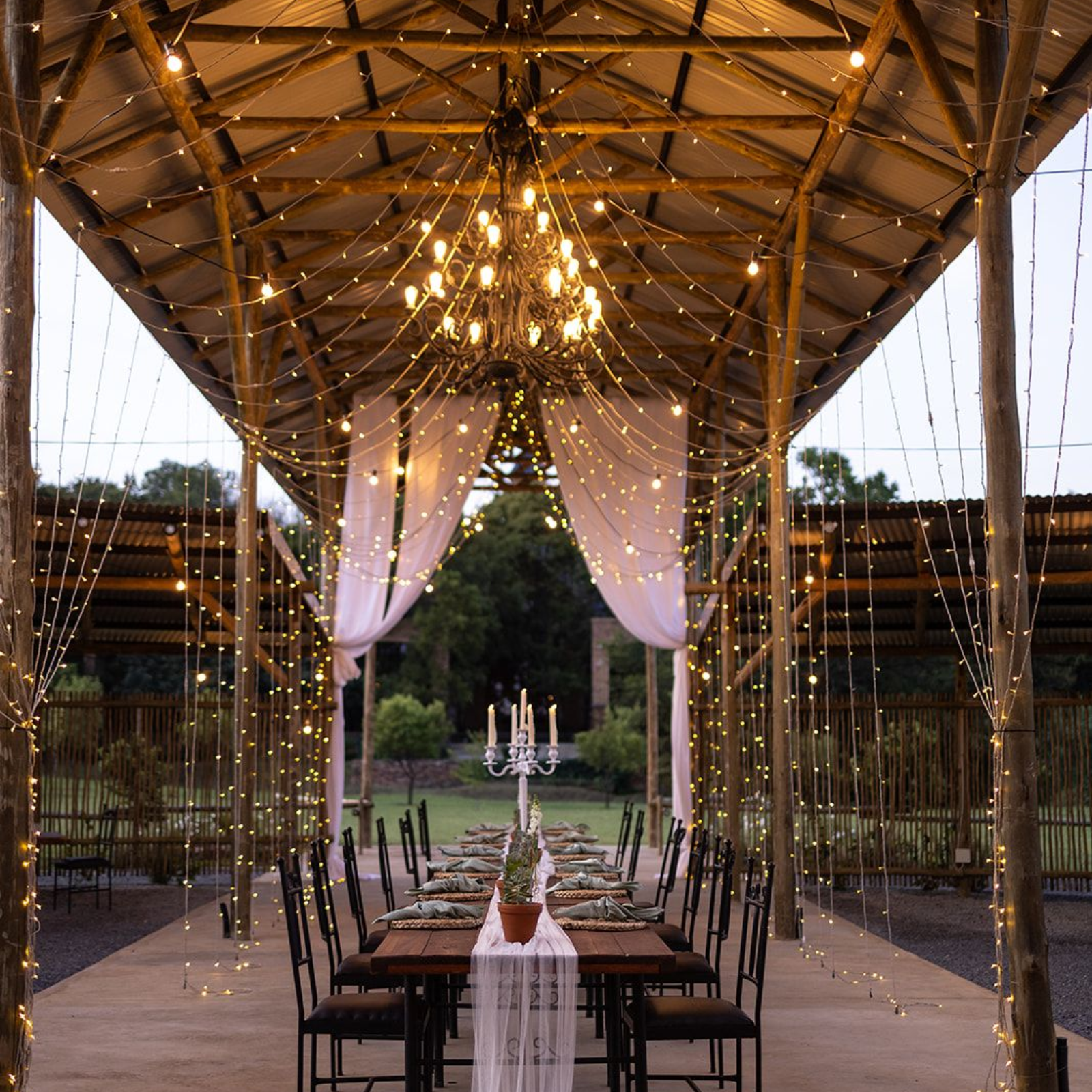 Elegant outdoor dining setup under a lit wooden pavilion with draped fabric and hanging lights