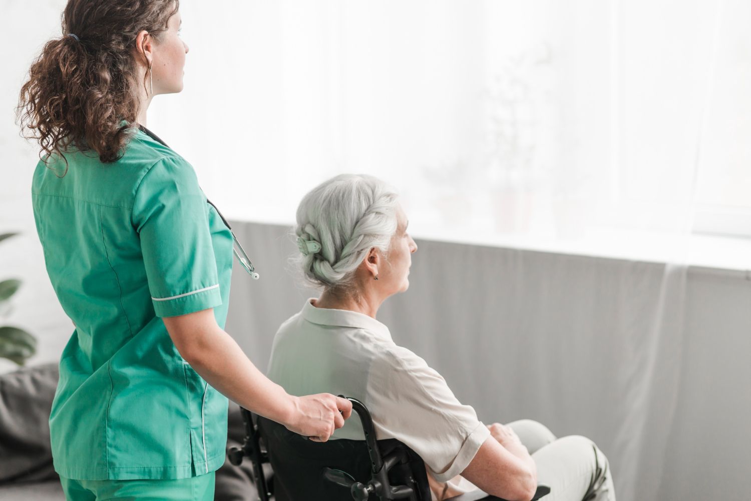 Nurse in green scrubs pushing a woman in a wheelchair toward a window.