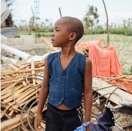 Boy in blue vest stands in debris-filled area, looking off to the side.