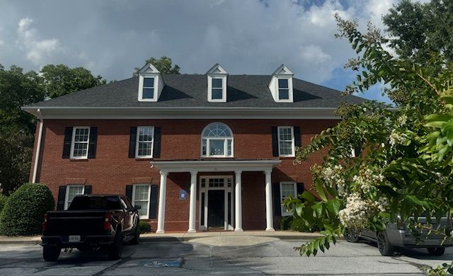 Two-story brick building with white columns at the entrance, black shutters, and three dormer windows under a dark roof.