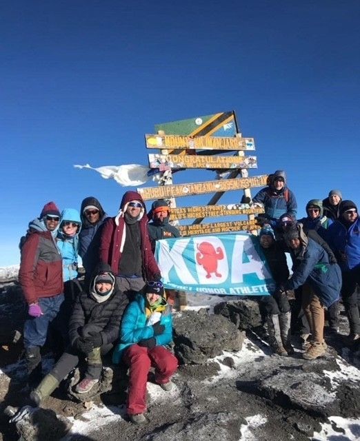 Group of people at Kilimanjaro summit, posing near wooden sign, blue sky, some snow, celebrating.