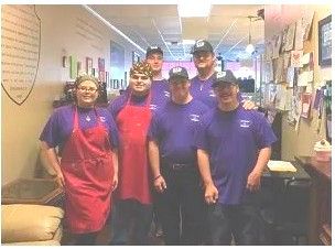 Group of six people in purple shirts and aprons stand in a hallway.