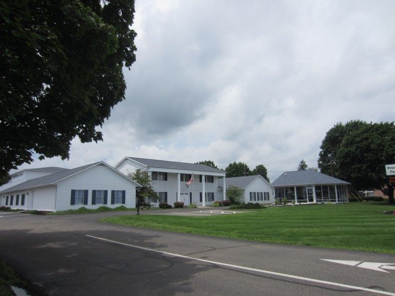 White buildings and a gazebo beside a paved road under a cloudy sky, with a green lawn in front