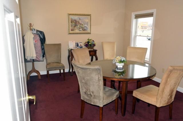 Dining room with round glass table, beige chairs, maroon carpet, and a coat rack by the wall.