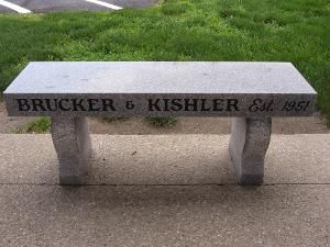 Stone bench engraved “Brucker & Kishler Est. 1951” beside a sidewalk and grass
