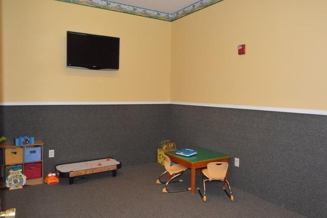 Quiet playroom with a wall-mounted TV, low table, chairs, and toys on a gray carpet.