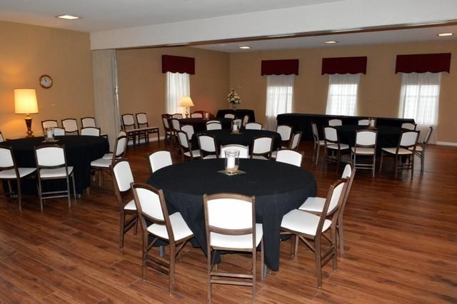 Empty banquet hall with round tables, black-and-white chairs, wood floors, and red window valances