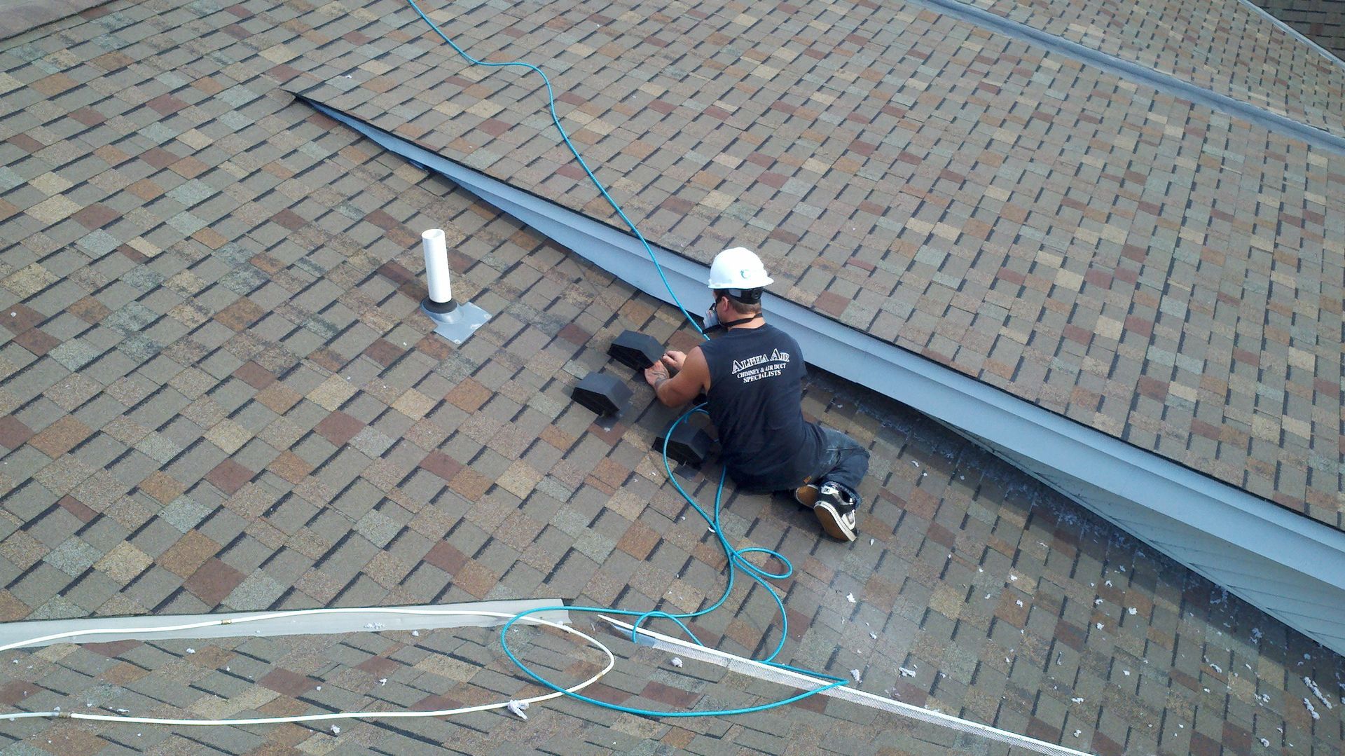 Roofer in hardhat working on a roof, cutting flashing. Brown and tan shingles, gray metal flashing, blue safety rope.