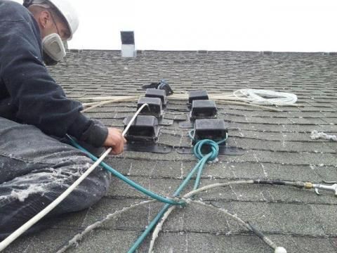 Roofer in safety gear, installing equipment on a shingle roof with ropes and black blocks.