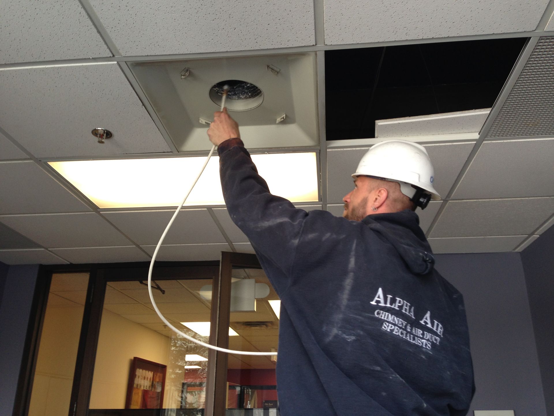 Man in hard hat inspecting ceiling vent with a probe in an office setting.