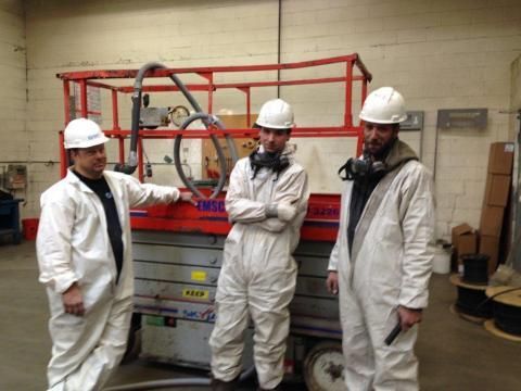 Three workers in white coveralls and hard hats stand near a scissor lift in a warehouse.