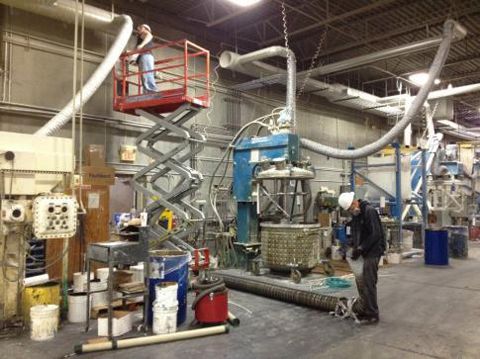 Two workers near industrial machinery, one in a lift, one on the ground. Interior of a factory.