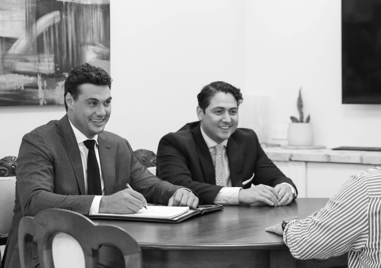 Two men in suits sit at a table, smiling and looking toward a person out of frame.