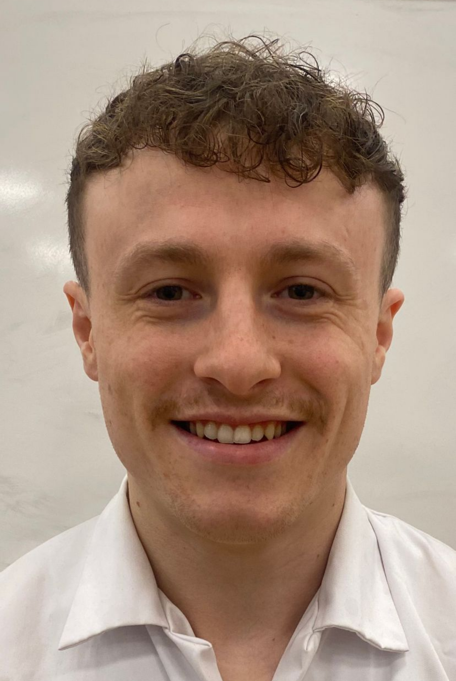 A person with short, curly brown hair and a light mustache, smiling directly at the camera while wearing a white shirt.