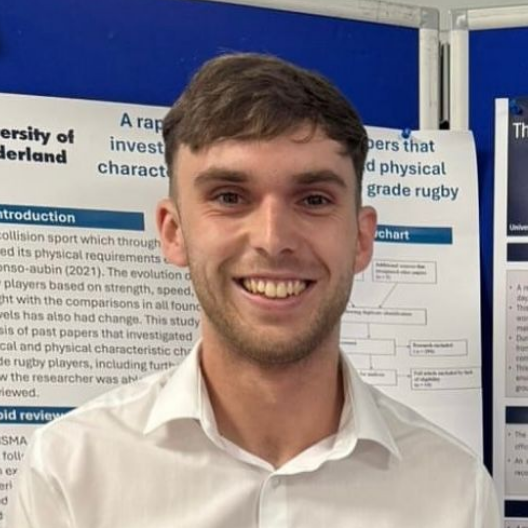 A smiling person in a white collared shirt stands in front of a research poster board at an academic presentation.
