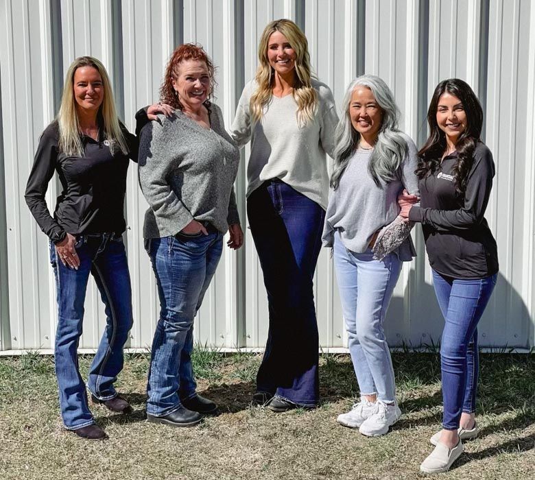 Five women posing together in front of a white building; jeans, casual tops, some smiling, arm around shoulders.