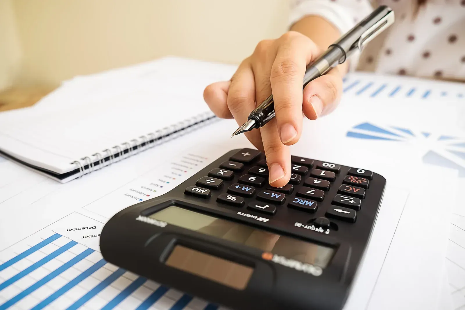 Person's hand using a calculator, holding a pen. Documents and a notepad are nearby.