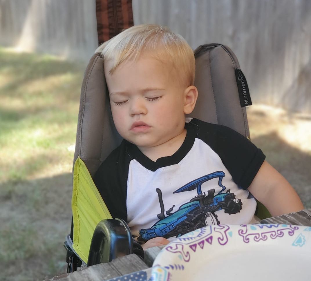 A little boy is sitting in a high chair with his eyes closed.