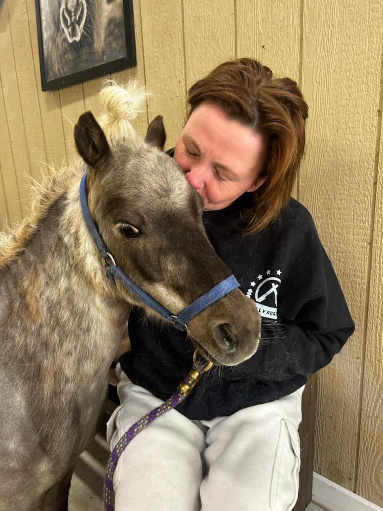 Person cuddling a small brown horse in a stable, both wearing blue halters.