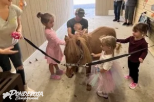 Children petting a small pony indoors, with adults nearby holding the lead rope